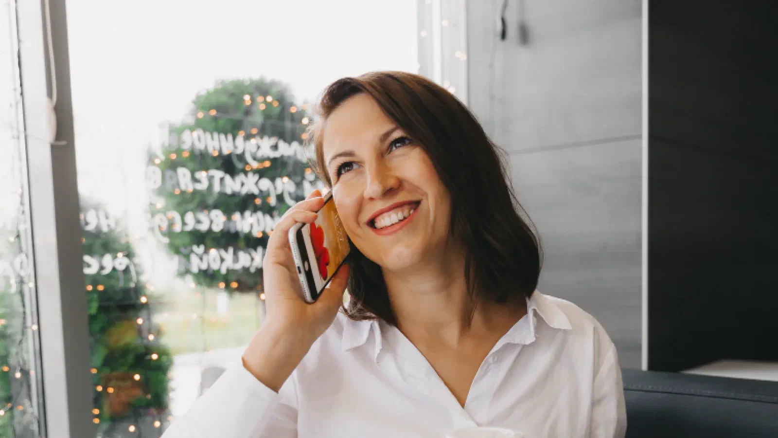 Mulher sorridente falando ao celular, com um copo de café, perto de uma janela
